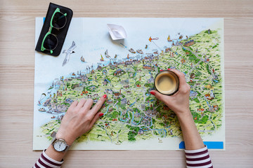 the girl's hand holds a cup of coffee and with the other hand indicates the terrain on the map where to go on a trip, on the table is a unfolded map on the map a paper white boat, top view, horizontal