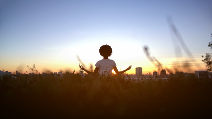 Young woman practicing yoga on hill at sunset, sitting in lotus pose, meditating