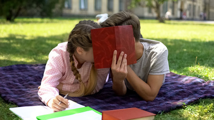 Teens hiding behind book and kissing, lying on plaid in park, first relations