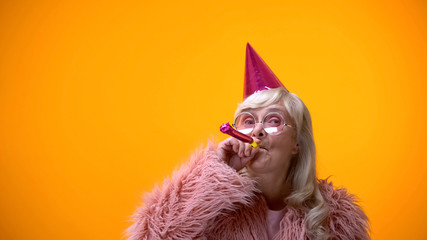 Glamorous aged woman blowing party horn, celebrating birthday anniversary