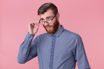Photo of a young attractive bearded man, looking disapprovingly through his glasses, colleague made a stupid mistake in work. Isolated over pink background.