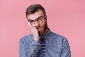 Portrait of young bearded businessman wears glasses feels unhappy and tired, propping up his head,...
