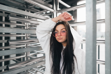 Street portrait of an attractive girl in street clothes near turnstiles. Stylish girl model in pink glasses and a white jacket poses for a futuristic location. Street fashion concept.