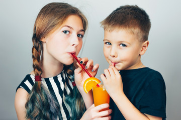 Beautiful girl and pretty little boy drinking orange fresh juice on gray studio background.