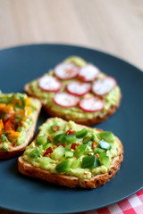 Three slices of toast with mashed avocado and various vegetable and herb toppings. Selective focus.