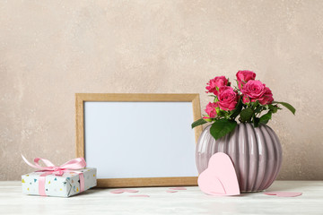 Vase with pink roses, empty frame, gift and little hearts on white table against light brown background