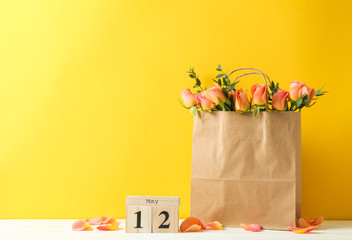 Paper bag with bouquet of orange roses and calendar on white table against color background, space for text