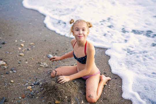 Having fun and joy concept. Blonde lovely girl with two braids playing with sea sand. Playful active kid on beach with waves in summer vacation