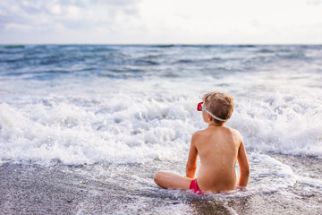 Child boy playing in the waves on the beach in summer sunset, kid watching sea waves and having fun