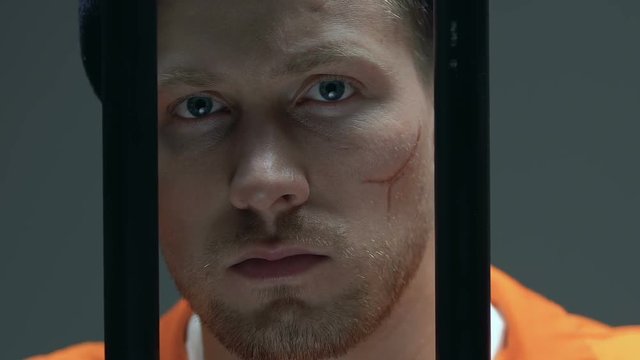 Desperate prisoner with scars on face looking through cell bars, incarceration
