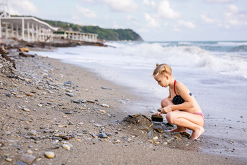 Having fun and joy concept. Blonde lovely girl with two braids playing with sea sand. Playful active kid on beach with waves in summer vacation