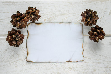On white wooden boards, a sheet of paper burned along the edges, forest cones around the edges. Leaving space for text.