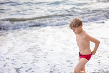 Blonde boy running and jumping on the beach on blue sea shore in summer vacation at the day time. Blue ocean with white big wawes on the background
