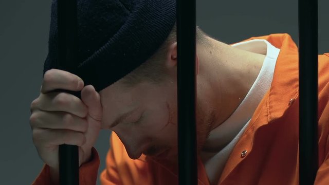 Upset prisoner with scars on face holding cell bars, regretting about made crime