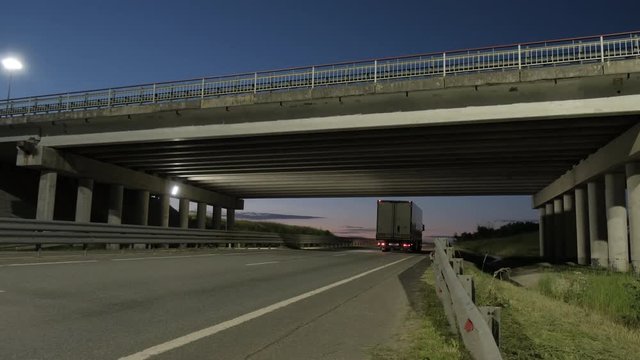 Cars and trucks on the highway under the bridge and on the bridge. View of the road from under the bridge. Suburban car traffic on the road at dusk. Sunset in the background. Blue and pink sky.