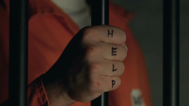 Prisoner with help word on beaten fists holding cell bars, aggression in jail