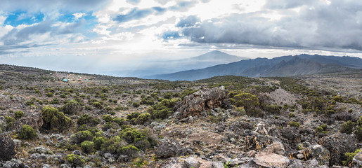 Fototapeta na wymiar Panoramic view of the Shira Cave Camp site on the Machame hiking route on Mt Kilimanjaro, Tanzania. Mount Meru is behind sun rays in the background under a dramatic cloudy sky, with rocks and foliage.
