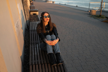 Young woman listens to music in closed headphones through her phone wearing a leather jacket and jeans at a sunset near river Daugava