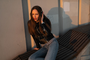 Young woman listens to music in closed headphones through her phone wearing a leather jacket and jeans at a sunset near river Daugava