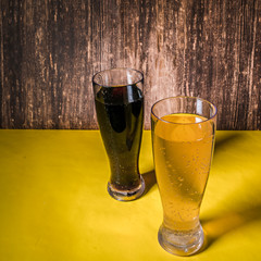 Two beers on a yellow bar next to a wooden walll.