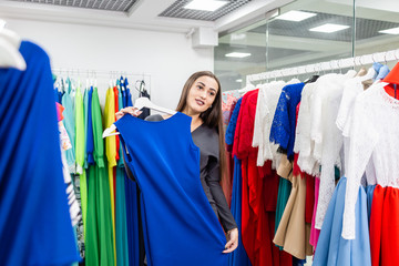 Naklejka na ściany i meble Beautiful girl with blue dress near mirror on room background. Happy young woman choosing clothes in mall or clothing store. Sale, fashion, consumerism concept