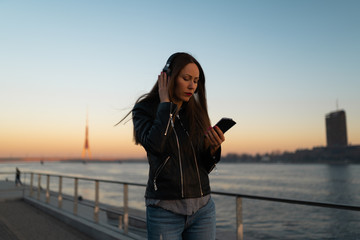 Young woman listens to music in closed headphones through her phone wearing a leather jacket and jeans at a sunset near river Daugava