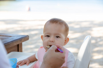 Baby girl eating porridge. Dad feeds little daughter with a spoon. Against the backdrop of a tropical beach.