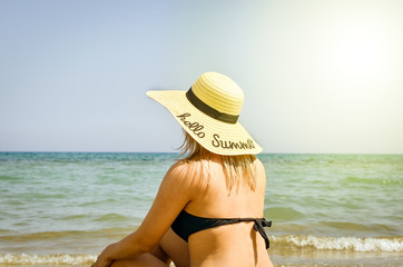 Fototapeta na wymiar Blonde girl dressed in a black swimsuits and big straw hat on his head which is tanning and relaxing to the seashore ..Bright warm filter .