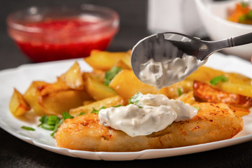 Fried fish and chips on a white plate on the kitchen table with tomato sauce, tartar sauce. Inpose...