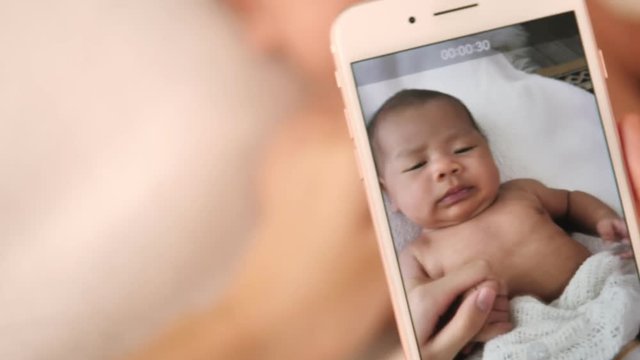 Mother recording video her newborn baby in bed with smart phone at home.