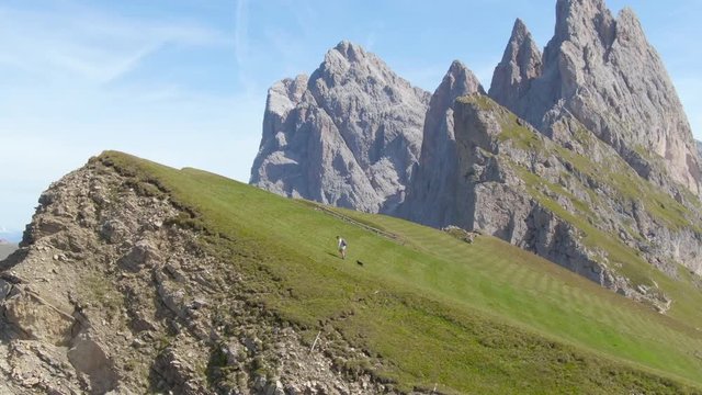 AERIAL: Flying next to a young woman hiking up the grassy hill in the Dolomites with her puppy. Female hiker treks up towards the mountaintop in the picturesque Italian Alps on a warm spring day.