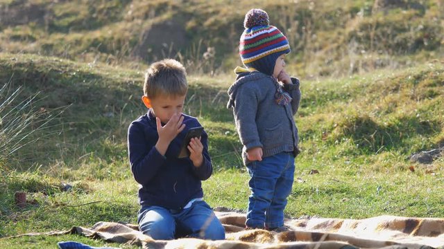 Biggest boy look at phone, baby child brother standing and look around in nature, technology trap and and joy of innocent childhood