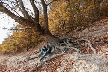 Redhead man in a plaid shirt and a purple t-shirt with backpack sits under a tilted tree in the midst of roots and fallen leaves in the autumn forest.