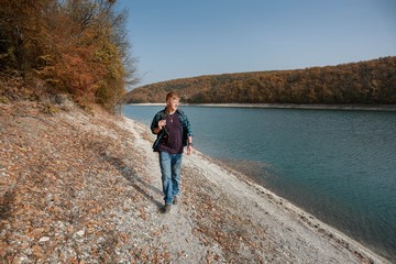 Redhead man in a plaid shirt and a purple t-shirt with backpack walks near the lake in the autumn forest