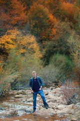 Naklejka premium Redhead man in a plaid shirt and purple t-shirt stands on the stones in the mountain stream in the autumn forest in the Grand Canyon of Crimea. Autumn, travel and hiking concept.