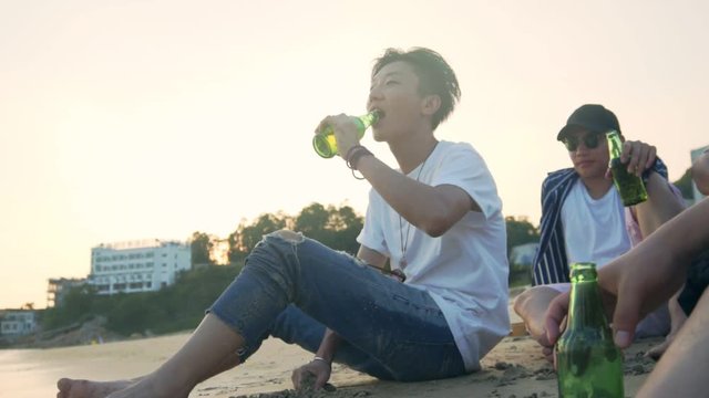 young asian adults sitting relaxing on beach drinking beer, slow motion.