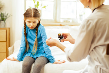 Small sick girl dissatisfiedly looking at pediatrician pouring throat syrup