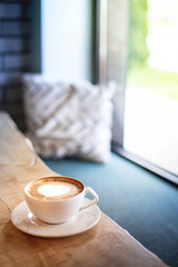 Morning scene with white cappuccino cup placed on light brown wood table next to window