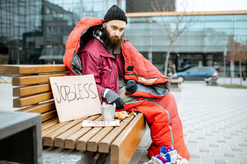 Homeless and jobless beggar sitting on the bench wrapped with sleeping bag begging money near the...