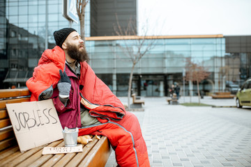 Homeless and jobless beggar sitting on the bench wrapped with sleeping bag begging money near the...