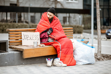 Homeless and jobless beggar sitting on the bench wrapped with sleeping bag begging money near the...
