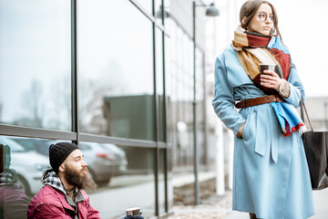 Depressed homeless begging money while sitting on the sidewalk with passing by businesswoman near...