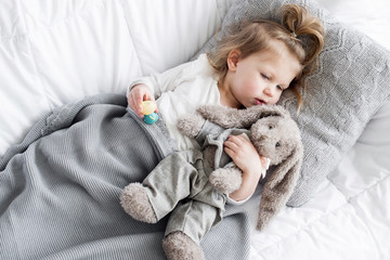 A little girl lies in the bed and embracing a toy hare. She is covered with a gray knitted blanket.