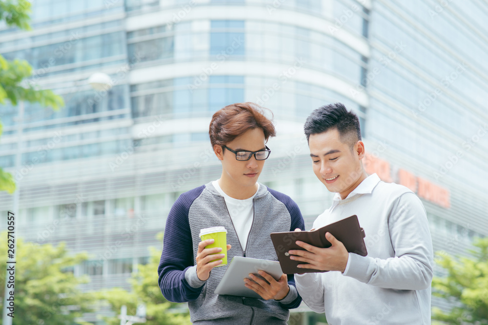 Wall mural Closeup of two smiling business men using tablet computer and walking with office building in background - Image