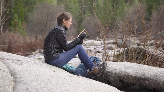 Beautiful woman reading on rock near a river after a hike
