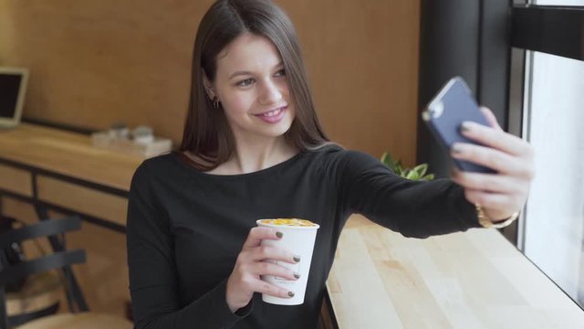 Close-up of a girl with beautiful smile and cute face is drinking coffee in cafe and taking selfies. Lifestyle.