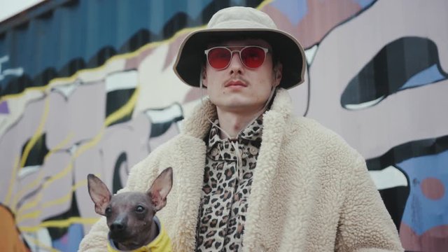 Portrait young man with hat in red sunglasses holding dog wear yellow clother in arms look at camera serious fashion style background artwall fun pet small young adult expression slow motion