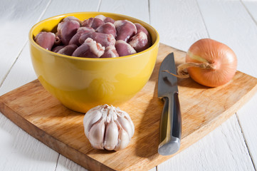 Chicken hearts in a yellow bowl on a cutting board. Near the bulb of garlic, one onion and a knife. Useful diet food.