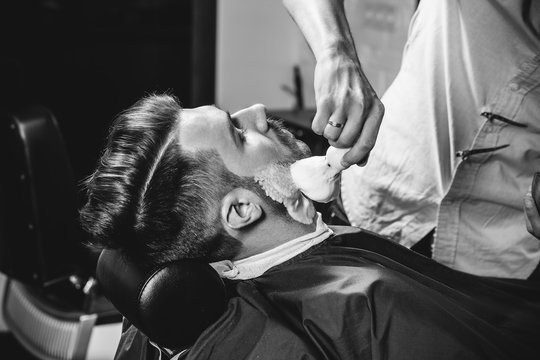 Client during beard shaving in barbershop. Young handsome barber and attractive bearded man. Black and white or colorless photo. Hairstyle, salon, hairdresser, barber shop, lifestyle concept