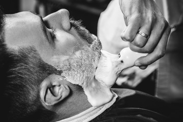 Client during beard shaving in barbershop. Young handsome barber and attractive bearded man. Black and white or colorless photo. Hairstyle, salon, hairdresser, barber shop, lifestyle concept